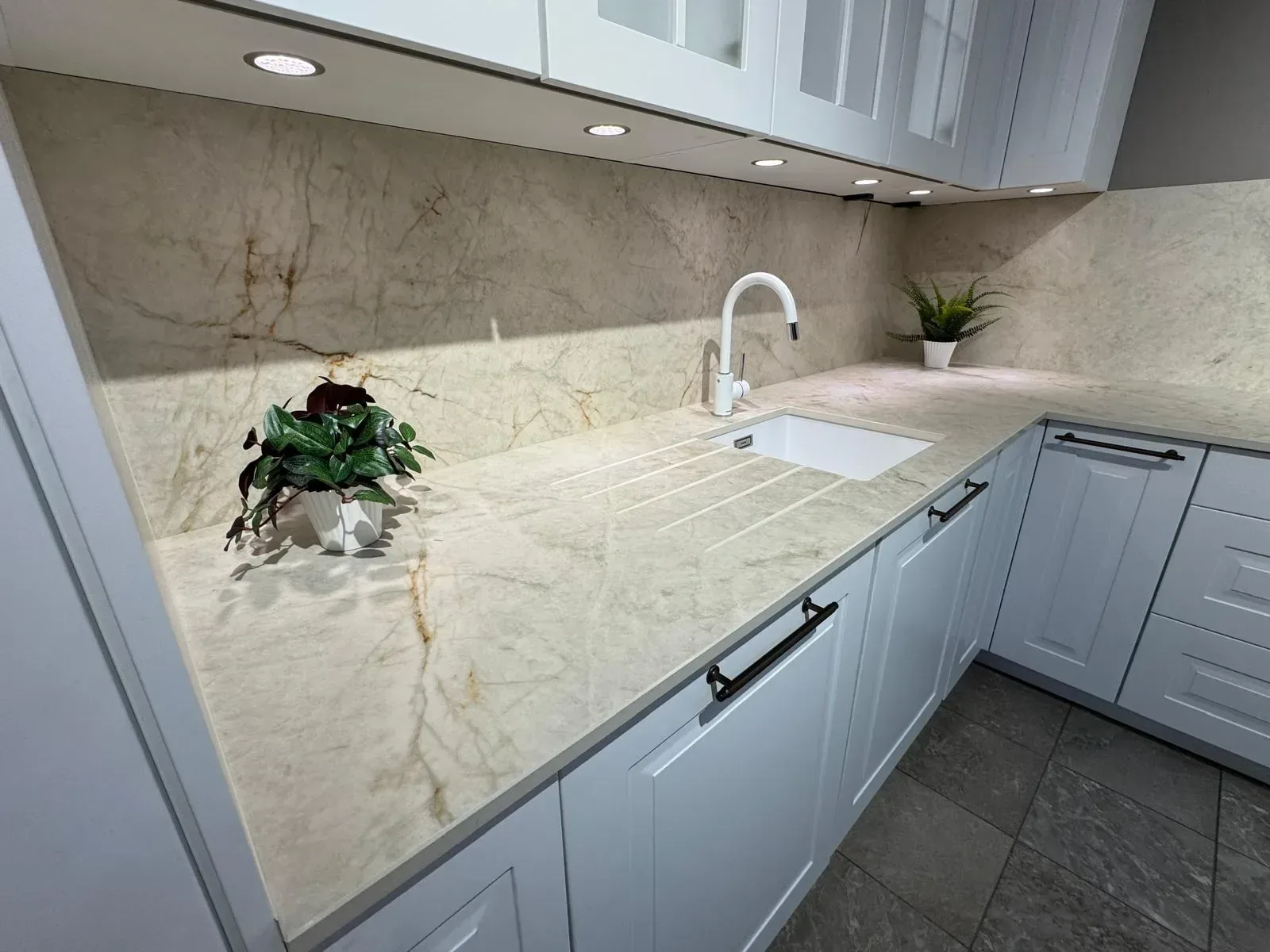Cuisine blanche avec plan de travail d’angle et crédence en pierre veinée beige, évier blanc sous-plan et rainures d’égouttoir