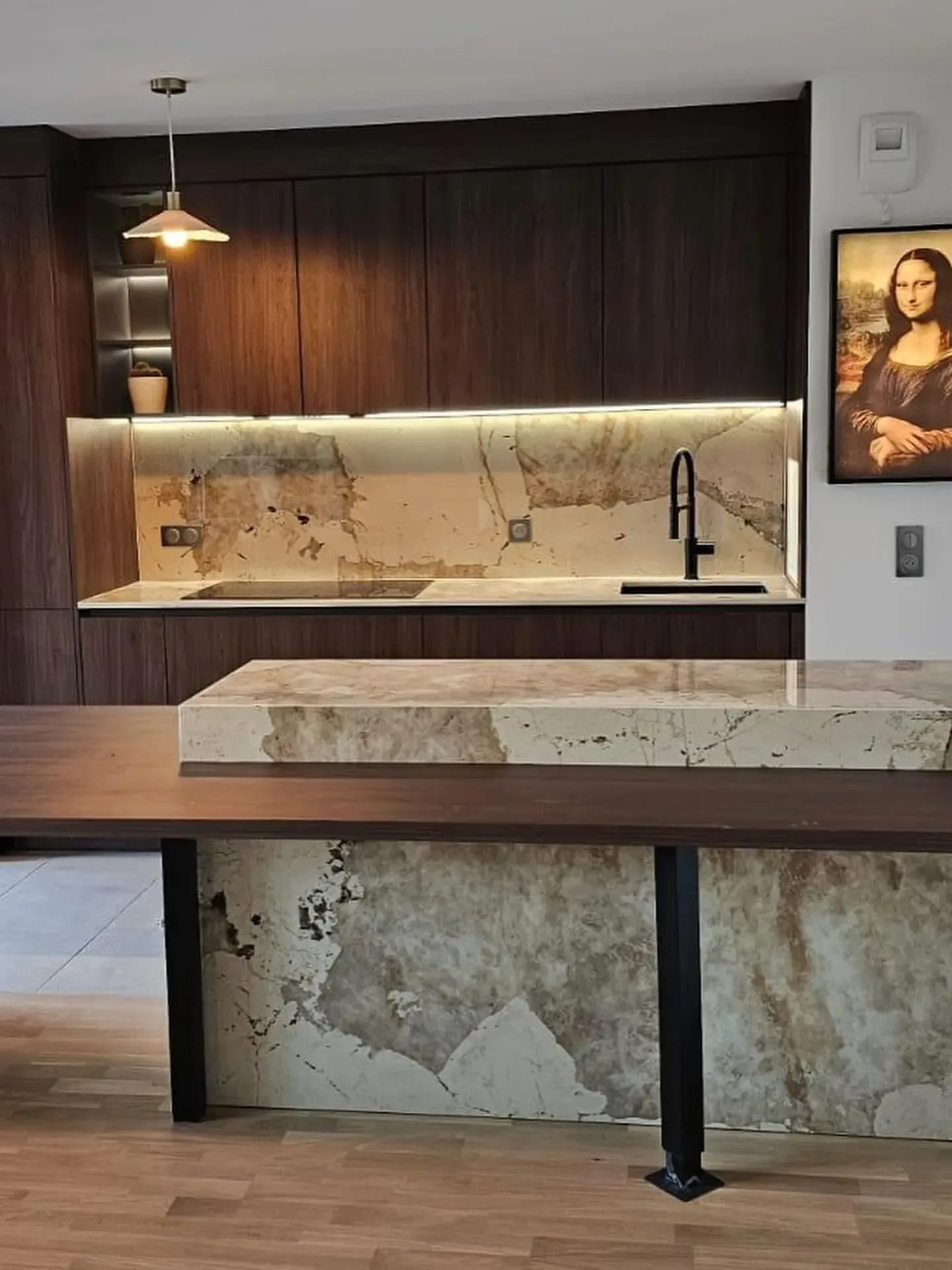 Cuisine en bois foncé avec crédence en pierre beige éclairée par un bandeau lumineux, et îlot habillé de la même pierre prolongé par une table en bois